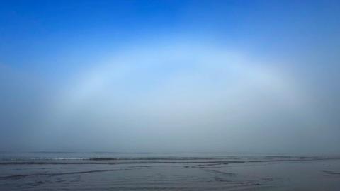 A fog bow in a bright blue sky over a very flat beach