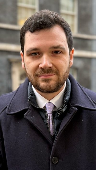 A man, Henry Zeffman, with short brown hair and a dark blue jacket, stands outside 10 Downing Street